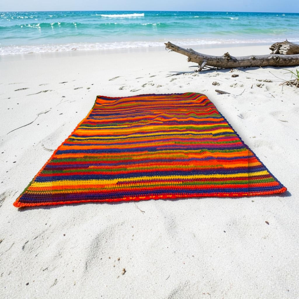 Rainbow stripe knit blanket laid out on white beach sand at the water's edge.