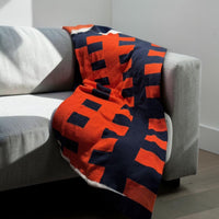 Knit throw in a bold orange and navy interlocking colorblock pattern, draped over a grey sofa.